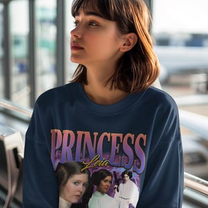 May include: A navy blue sweatshirt with a vintage-style graphic print of Princess Leia from Star Wars. The graphic features a collage of images of Princess Leia in different poses, with the text "Princess Leia" in a colorful, gradient font.
