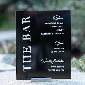 May include: Black acrylic sign with white text that reads "THE BAR" and lists drink options: Wine, Sauvignon Blanc, Pinot Gris, Merlot, Cabernet Sauvignon, Beer, Local IPA, Local Lager, Non Alcoholic, Soft Drinks, Coffee and Tea.