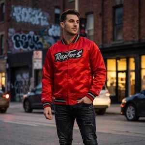 May include: A bright red satin bomber jacket featuring the word Raptors in black and white lettering across the chest. The jacket has a snap-button front closure, long sleeves, and ribbed cuffs and collar with black, white, and red striped detailing.
