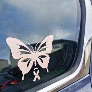May include: A pale pink butterfly sticker with a ribbon symbol, attached to a car window. The butterfly has intricate wing patterns. The ribbon is a symbol of awareness. The sticker is a decorative item.