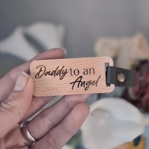 May include: A rectangular wooden tag with the engraved text "Daddy to an Angel". The tag has a dark gray leather strap attached with a metal rivet. The background is blurred, with white flowers visible.
