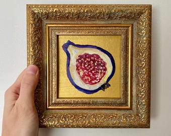 May include: A framed painting of a fig. The fig is cut in half, revealing a red interior with small seeds, surrounded by a white and blue outline. The painting is set against a gold background and framed in an ornate gold frame.
