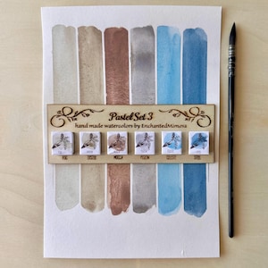 Peut inclure: Un ensemble de peinture aquarelle intitulé "Pastel Set 3" avec six échantillons de couleurs dans des tons neutres et bleus. L'ensemble comprend un support en bois avec la marque "EnchantedMimosa" et un pinceau.
