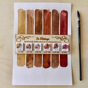 Op de afbeelding: Een aquarelverfset met het label "So Vintage" met verschillende tinten bruin en rood. De set bevat een houten houder met de namen van de kleuren, zoals "Clear Sienna" en "Red Cherry". Rechts van de set is een penseel geplaatst.