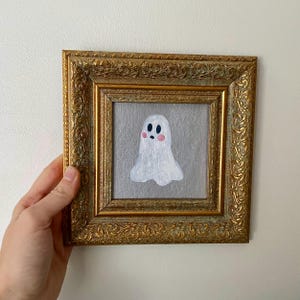 Può includere: Dipinto incorniciato di un fantasma bianco con guance rosa e occhi neri. L'opera d'arte è ambientata su uno sfondo grigio e incorniciata in una cornice dorata ornata. Il dipinto è tenuto da una mano.