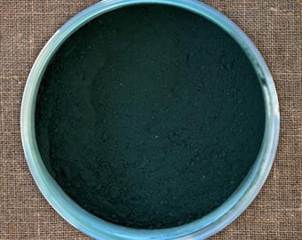 Puede incluir: Primer plano de un tarro de vidrio redondo lleno de un polvo verde oscuro. El polvo es liso y uniforme en textura.