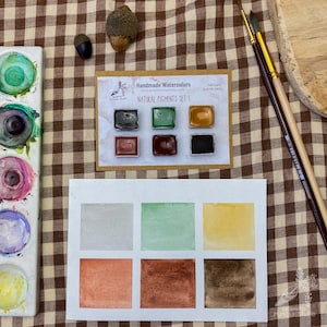May include: A set of six watercolour paints in small metal pans. The pans are labelled "Natural Pigments Set 1" and include the colours grey, green, yellow, orange, brown, and black. The paints are arranged on a brown and white checked fabric background.