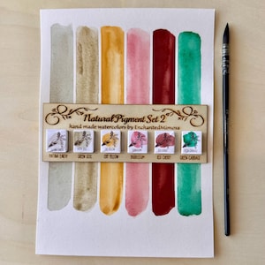 Op de afbeelding: Een aquarelset met het label "Natural Pigment Set 2" met kleurstalen van Patina Earth, Green Soil, Oat Yellow, Bubblegum, Red Cherry en Green Cabbage. Een penseel staat rechts. Handgemaakt door EnchantedMimosa.