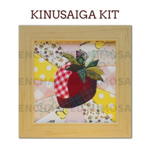 May include: A Kinusaiga kit featuring a square light wood frame containing a fabric mosaic of a strawberry. The central strawberry is composed of red gingham, plaid, and solid fabric pieces with green leaves, surrounded by yellow, floral, and patterned fabric rays.