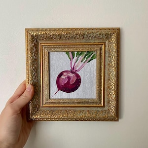 Puede incluir: Pintura enmarcada de una remolacha. La obra presenta una remolacha morada vibrante con hojas verdes y tallos rosas, sobre un fondo plateado. La pintura está encerrada en un marco dorado y ornamentado, agregando un toque de elegancia.