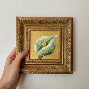 Può includere: Piccolo dipinto quadrato di un bocciolo di calla bianco e verde con centro giallo. L'opera d'arte è incorniciata in una cornice dorata ornata. Il dipinto è presentato su uno sfondo dorato. Una mano tiene il dipinto.