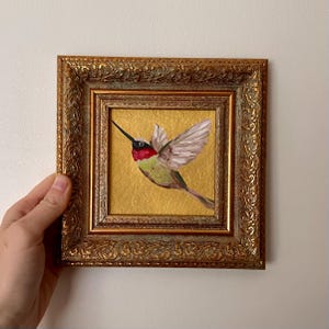 Op de afbeelding: Een ingelijste schildering van een kolibrie in vlucht. De vogel heeft een rode keel, een groen lichaam en witte vleugels. De schildering staat tegen een gouden achtergrond en is ingelijst in een sierlijke gouden lijst.