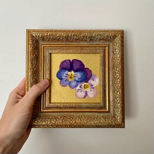 以下が含まれることがあります： 2つのパンジーの額入り絵画。パンジーは紫、青、白の色合いで描かれ、金色の背景に配置されています。フレームは装飾的で、金色の仕上げが施されています。このアートワークは手で持たれています。