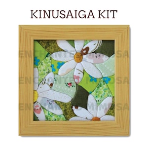 Pode incluir: Um kit Kinusaiga quadrado apresentando um mosaico de tecido de margaridas brancas com centros amarelos sobre um fundo verde, tudo dentro de uma moldura de madeira clara. O texto KINUSAIGA KIT está impresso em castanho sobre a obra emoldurada.