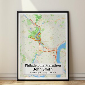 May include: A black and white map of Philadelphia with an orange line marking the route of the Philadelphia Marathon. The text "Philadelphia Marathon" and "John Smith" are printed below the map, along with the date "11/24/2024", the distance "26.2 Miles", and the time "04:12:16".