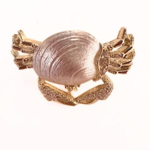 Peut inclure: Une broche en forme de crabe dorée avec une coquille rose et blanche. Les pinces du crabe sont ornées de petits strass.