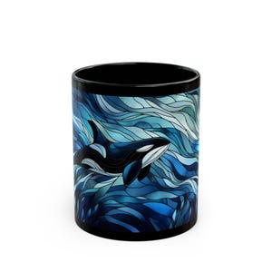 May include: Black coffee mug featuring a stylized illustration of a killer whale swimming in blue ocean waves. The orca is depicted in black and white, with the background showcasing shades of blue. The mug has a black rim and interior.