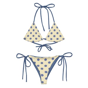 May include: A two-piece bikini set. The top is a halter style with triangle cups. The bottom is a tie-side style. Both pieces are cream-coloured with blue polka dots and blue trim.