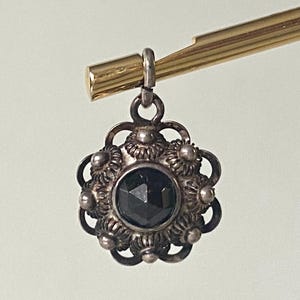 May include: A vintage silver pendant with a dark faceted gemstone center. The pendant has an ornate floral design around the gem and is suspended from a gold-colored metal bar.