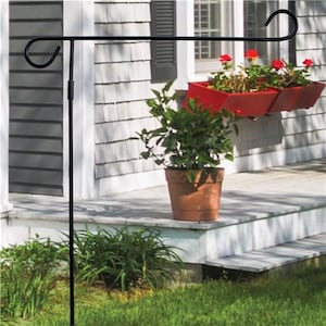 May include: Black metal garden flag stand with a curved arm. The stand is in front of a house with white siding and a window with red flowers in a planter.