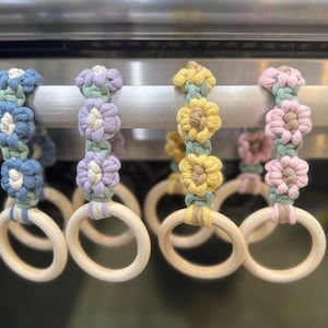 May include: Four macrame flower rings in blue, purple, yellow, and pink. Each ring has a wooden base and is adorned with a series of handmade macrame flowers. The flowers are attached to a wooden dowel.