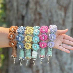 May include: A collection of colorful macrame flower wristlets in shades of orange, blue, yellow, turquoise, pink, and lavender. Each wristlet features a flower design and a silver-colored clasp.