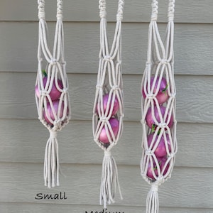 May include: Three macrame plant hangers in varying sizes, each filled with purple onions. The hangers are made of beige rope and feature a braided top loop and a tassel at the bottom. The sizes are labeled "Small", "Medium", and "Large".