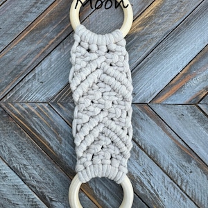 Pu&ograve; includere: Un arazzo in macram&egrave; con un design a corda annodata beige chiaro fissato a due anelli di legno circolari e lisci. La parola Moon &egrave; stampata in corsivo nero vicino all'anello di legno superiore, centrata sopra l'intricato motivo intrecciato.