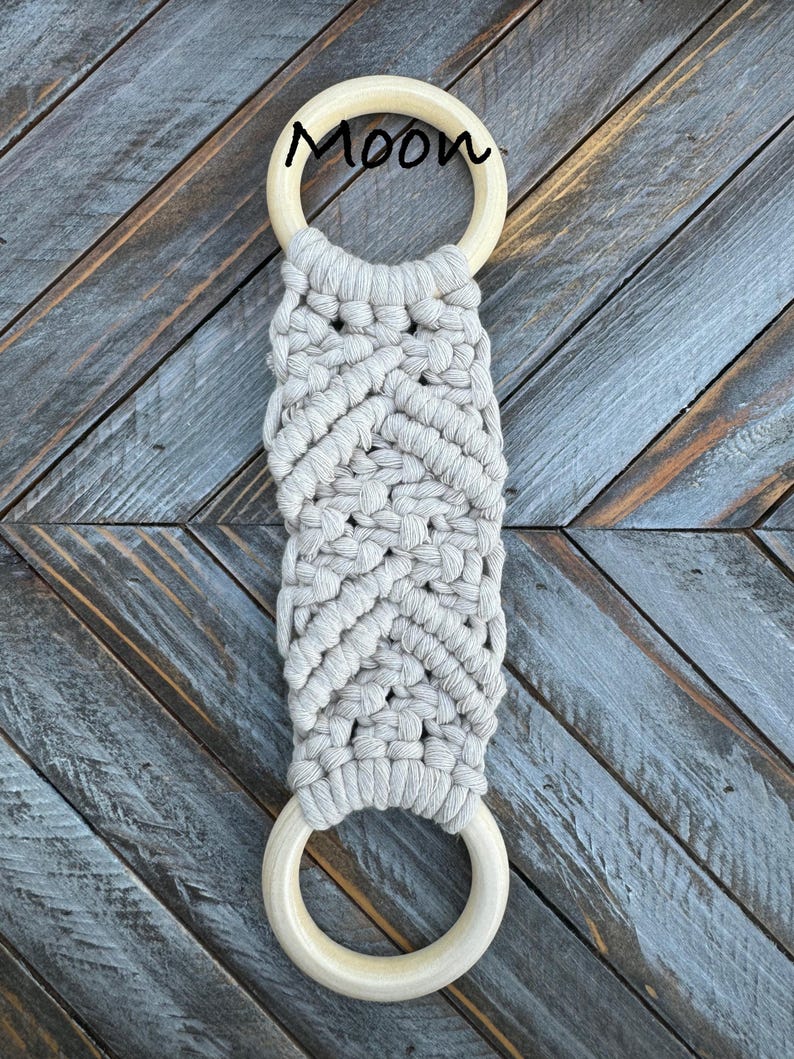 Pu&ograve; includere: Un arazzo in macram&egrave; con un design a corda annodata beige chiaro fissato a due anelli di legno circolari e lisci. La parola Moon &egrave; stampata in corsivo nero vicino all'anello di legno superiore, centrata sopra l'intricato motivo intrecciato.