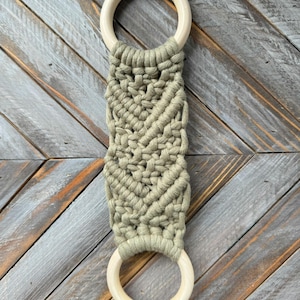 Pu&ograve; includere: Un accessorio in macram&egrave; con due anelli in legno chiaro e liscio collegati da una sezione centrale in corda di cotone verde salvia annodata con un motivo decorativo a spina di pesce. La parola Basil &egrave; stampata in nero sopra l'anello superiore.