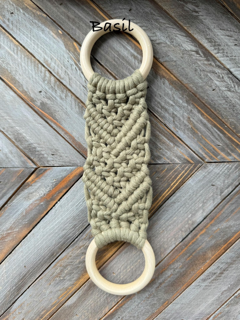 Pu&ograve; includere: Un accessorio in macram&egrave; con due anelli in legno chiaro e liscio collegati da una sezione centrale in corda di cotone verde salvia annodata con un motivo decorativo a spina di pesce. La parola Basil &egrave; stampata in nero sopra l'anello superiore.