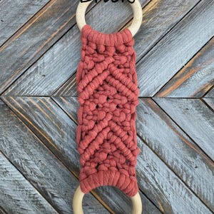 Pu&ograve; includere: Un arazzo in macram&egrave; con una corda color mattone annodata con un motivo a chevron tra due anelli di legno lisci e circolari. La parola Brick &egrave; stampata in nero sopra l'anello di legno superiore per indicare il colore della corda.