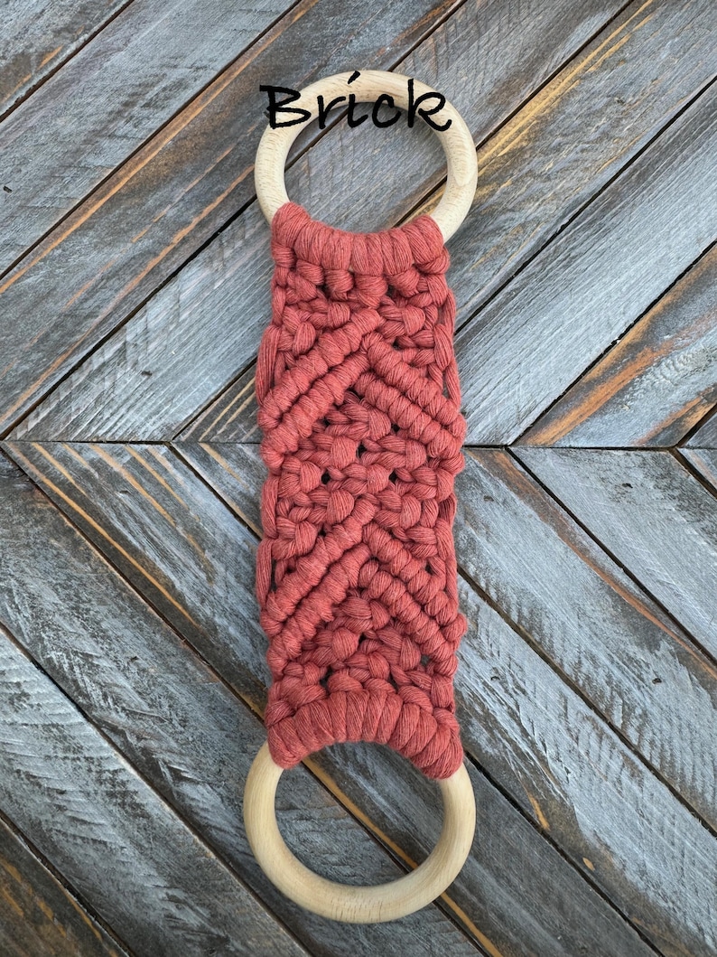 Pu&ograve; includere: Un arazzo in macram&egrave; con una corda color mattone annodata con un motivo a chevron tra due anelli di legno lisci e circolari. La parola Brick &egrave; stampata in nero sopra l'anello di legno superiore per indicare il colore della corda.