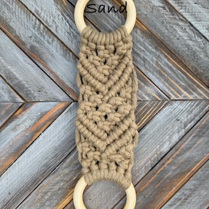 Pu&ograve; includere: Un arazzo in macram&egrave; con due anelli in legno chiaro e liscio collegati da una sezione centrale di corda color marrone chiaro annodata con un motivo a chevron. La parola Sand &egrave; stampata in nero vicino all'anello di legno superiore.