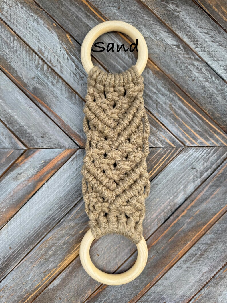 Pu&ograve; includere: Un arazzo in macram&egrave; con due anelli in legno chiaro e liscio collegati da una sezione centrale di corda color marrone chiaro annodata con un motivo a chevron. La parola Sand &egrave; stampata in nero vicino all'anello di legno superiore.