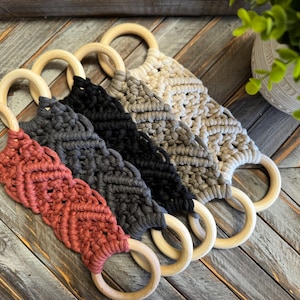 May include: Five macrame wall hangings with wooden rings in a row. The colors include rust, gray, black, gray-beige, and beige. The macrame has a diamond pattern. The wooden rings are light brown.