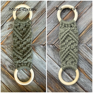 Pu&ograve; includere: Due portasciugamani in macram&egrave; con corda color verde muschio e foglia di alloro annodata a motivo chevron. Ogni pezzo ha un anello in legno chiaro e liscio nella parte superiore e inferiore. Il testo Moss Green e Bay Leaf &egrave; visibile sopra.