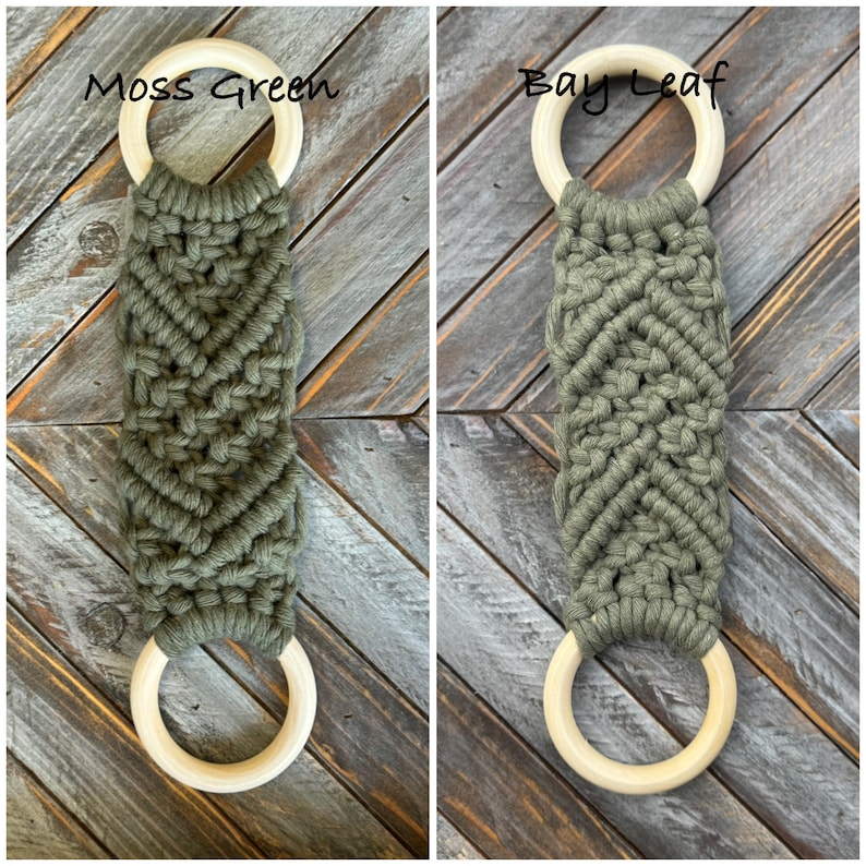 Pu&ograve; includere: Due portasciugamani in macram&egrave; con corda color verde muschio e foglia di alloro annodata a motivo chevron. Ogni pezzo ha un anello in legno chiaro e liscio nella parte superiore e inferiore. Il testo Moss Green e Bay Leaf &egrave; visibile sopra.
