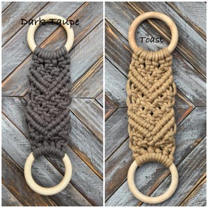 Pu&ograve; includere: Due arazzi in macram&egrave; con corda color tortora scuro e toast annodati con motivo a chevron. Ogni pezzo ha un anello in legno chiaro nella parte superiore e inferiore. Il testo Dark Taupe &egrave; scritto sopra il pezzo sinistro e Toast sopra quello destro.