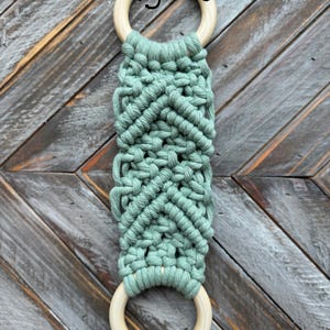Pu&ograve; includere: Un arazzo in macram&egrave; con corda verde salvia annodata a motivo chevron tra due anelli di legno chiaro e liscio. La parola Agave &egrave; stampata in nero vicino all'anello di legno superiore, mettendo in risalto un design strutturato.
