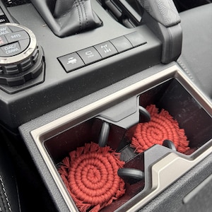 May include: Two red, circular, woven coasters with fringe sit in a car's center console cup holders. The coasters are made of a thick, textured material. The console is black with silver trim and buttons.