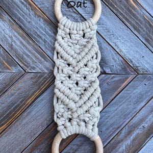 Pu&ograve; includere: Un portasciugamani in macram&egrave; con corda di cotone beige chiaro annodata in un motivo geometrico tra due anelli di legno chiaro, lisci e circolari. La parola Oat &egrave; stampata in nero vicino all'anello di legno superiore dell'oggetto.