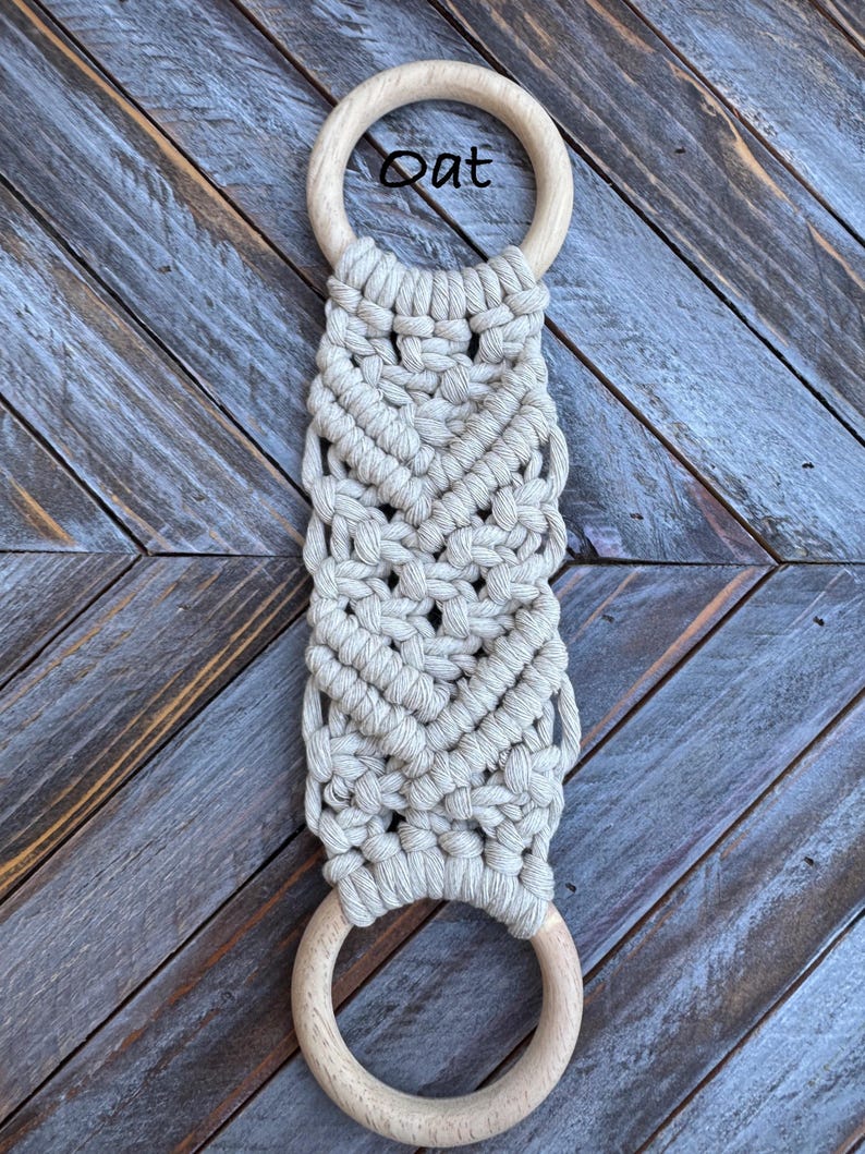 Pu&ograve; includere: Un portasciugamani in macram&egrave; con corda di cotone beige chiaro annodata in un motivo geometrico tra due anelli di legno chiaro, lisci e circolari. La parola Oat &egrave; stampata in nero vicino all'anello di legno superiore dell'oggetto.