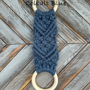Pu&ograve; includere: Un arazzo in macram&egrave; con corda blu scuro annodata a spina di pesce tra due anelli di legno lisci e chiari. Il testo Colbalt Blue &egrave; stampato in nero sopra l'anello di legno superiore sulla superficie strutturata.