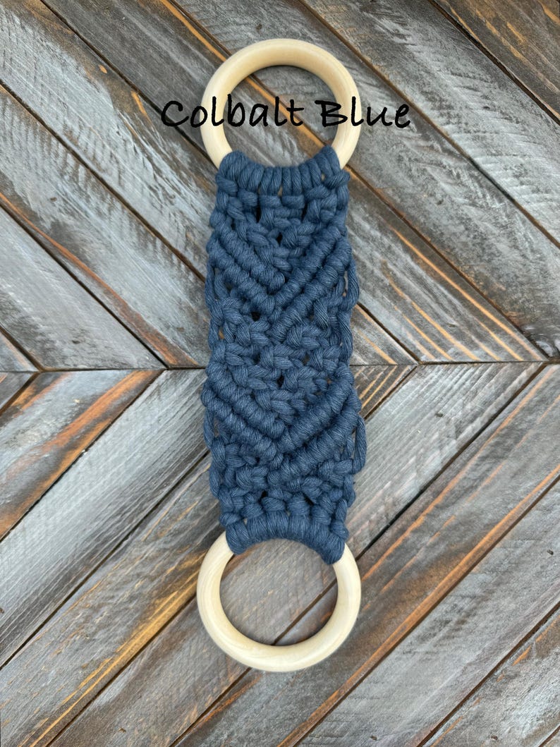 Pu&ograve; includere: Un arazzo in macram&egrave; con corda blu scuro annodata a spina di pesce tra due anelli di legno lisci e chiari. Il testo Colbalt Blue &egrave; stampato in nero sopra l'anello di legno superiore sulla superficie strutturata.