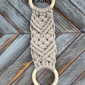 Pu&ograve; includere: Un portasciugamani in macram&egrave; con una corda color kaki annodata con un motivo a chevron tra due anelli di legno liscio di colore chiaro. La parola Khaki &egrave; stampata in nero vicino all'anello di legno superiore di questo accessorio.