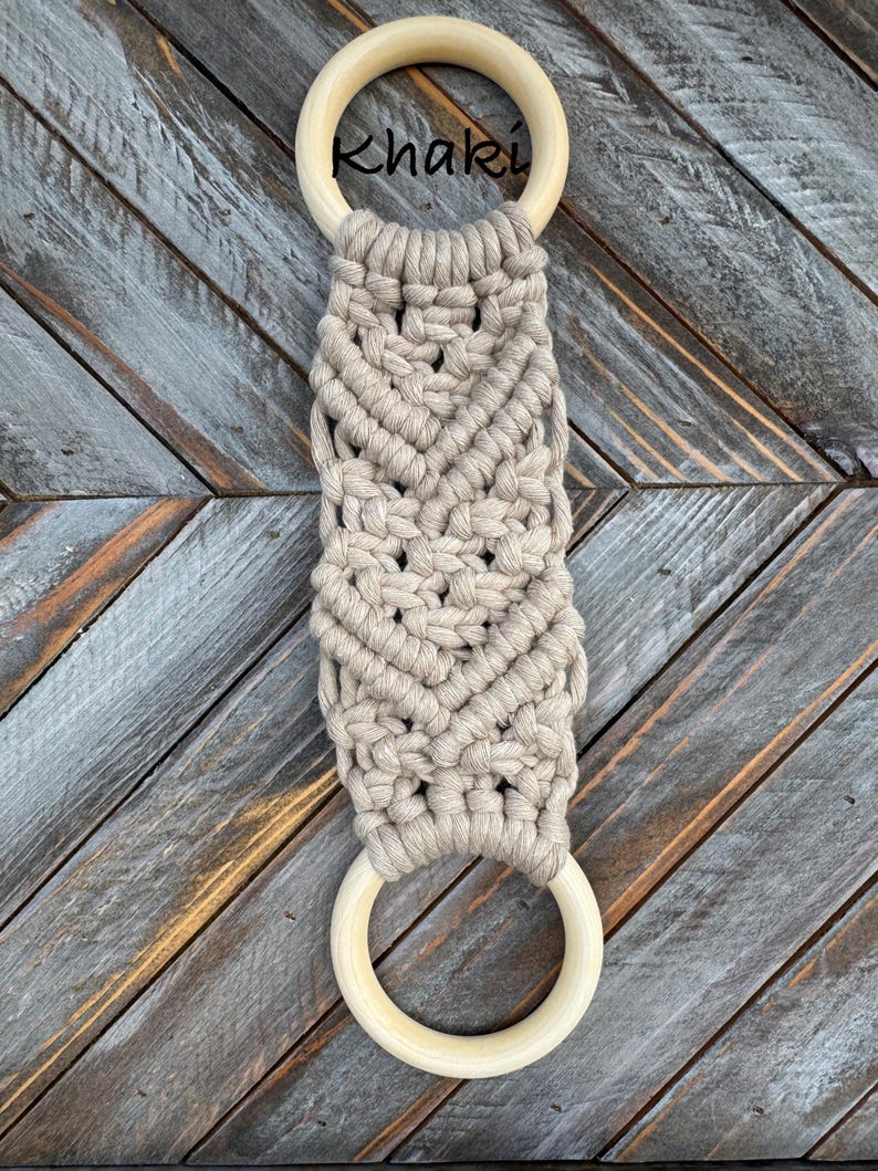 Pu&ograve; includere: Un portasciugamani in macram&egrave; con una corda color kaki annodata con un motivo a chevron tra due anelli di legno liscio di colore chiaro. La parola Khaki &egrave; stampata in nero vicino all'anello di legno superiore di questo accessorio.