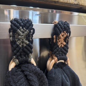 May include: Two black and white woven towel rings with wooden rings hanging on a stainless steel bar. One ring is black, white, and green, and the other is black, white, and brown.