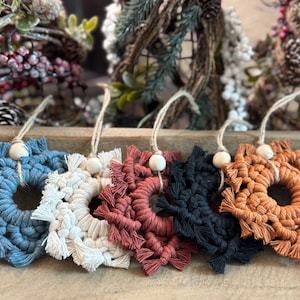 May include: A collection of five macrame snowflake ornaments in various colors: blue, white, red, black, and orange. Each ornament has a wooden bead and a natural twine loop for hanging. These decorative items are perfect for holiday decor.