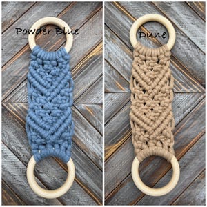 Pu&ograve; includere: Due portasciugamani in macram&egrave; con corda annodata nei colori blu polvere e duna. Ogni pezzo &egrave; costituito da una sezione centrale intrecciata fissata a due anelli di legno chiaro e liscio nella parte superiore e inferiore. Appare il testo Powder Blue e Dune.